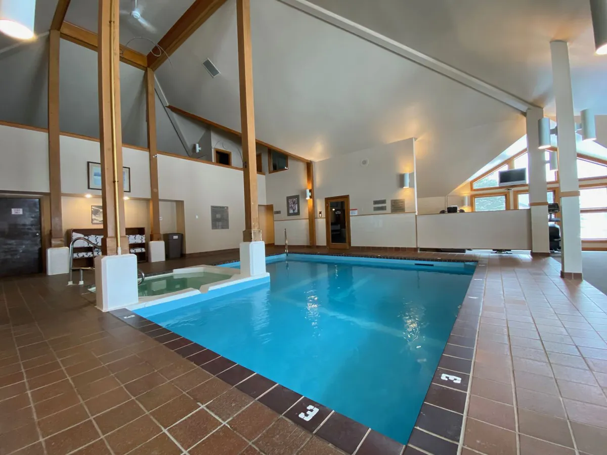The image features a spacious indoor swimming pool with large windows, wooden beams, and a lounge/reception area. It exudes a calm and relaxing vibe, ideal for family recreation regardless of the season.