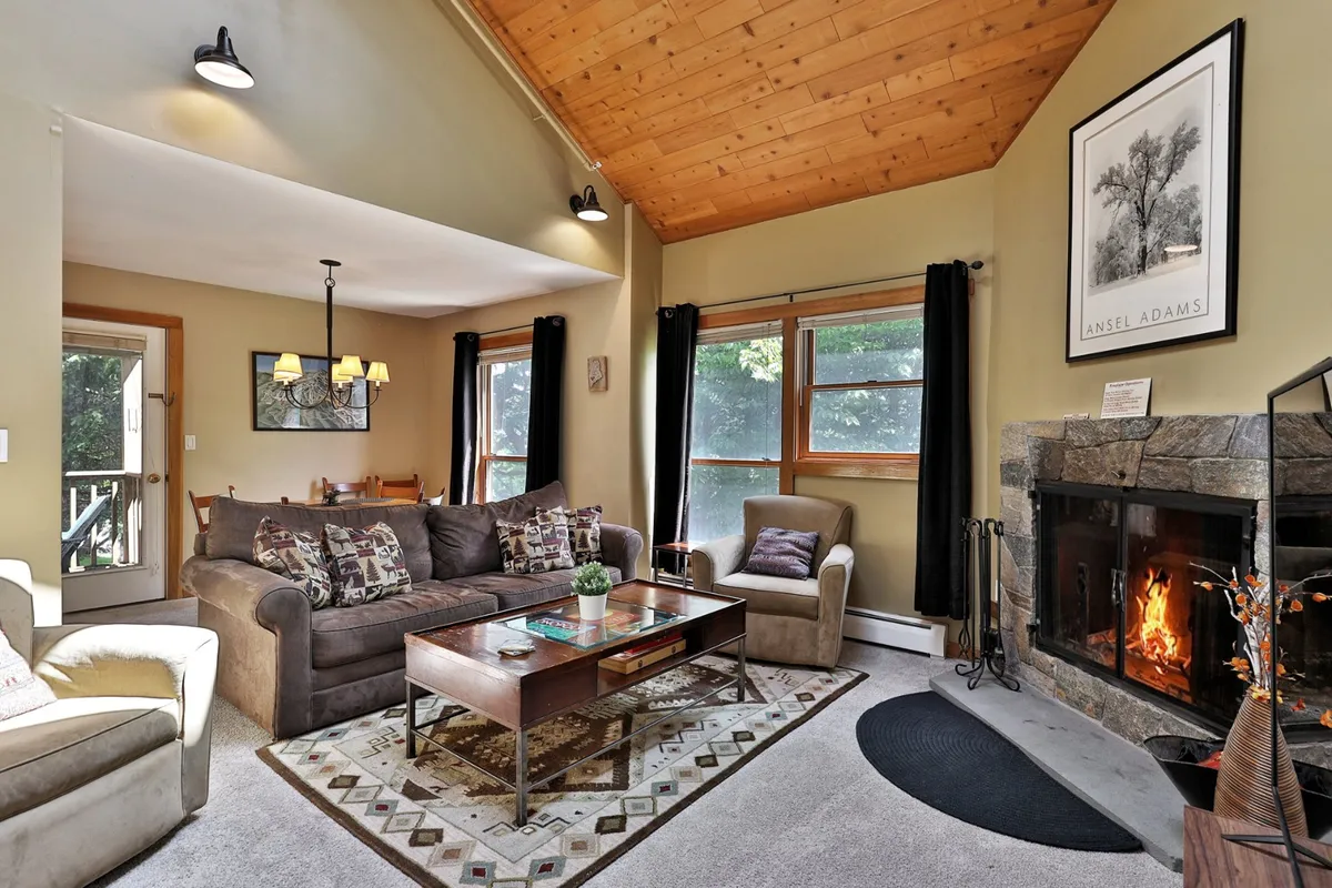 A warm, welcoming living room featuring a glowing wood-burning fireplace as its centerpiece. A comfortable sofa is positioned to enjoy both the fire and the expansive views through large windows, with tasteful artwork adding a homey touch. The ambiance is ideal for relaxing, sharing stories, or unwinding after a day outdoors.