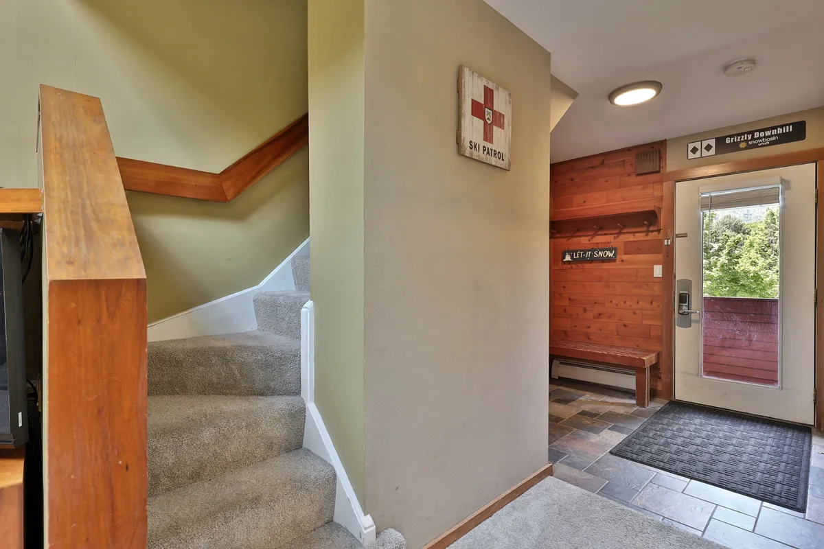 A welcoming entryway with a wooden bench, a staircase, and a vintage ski patrol sign sets the stage for gearing up before a day on the slopes or returning after outdoor adventures. The cozy, practical setup is designed for skiers and snowboarders to transition between the indoors and outdoors.