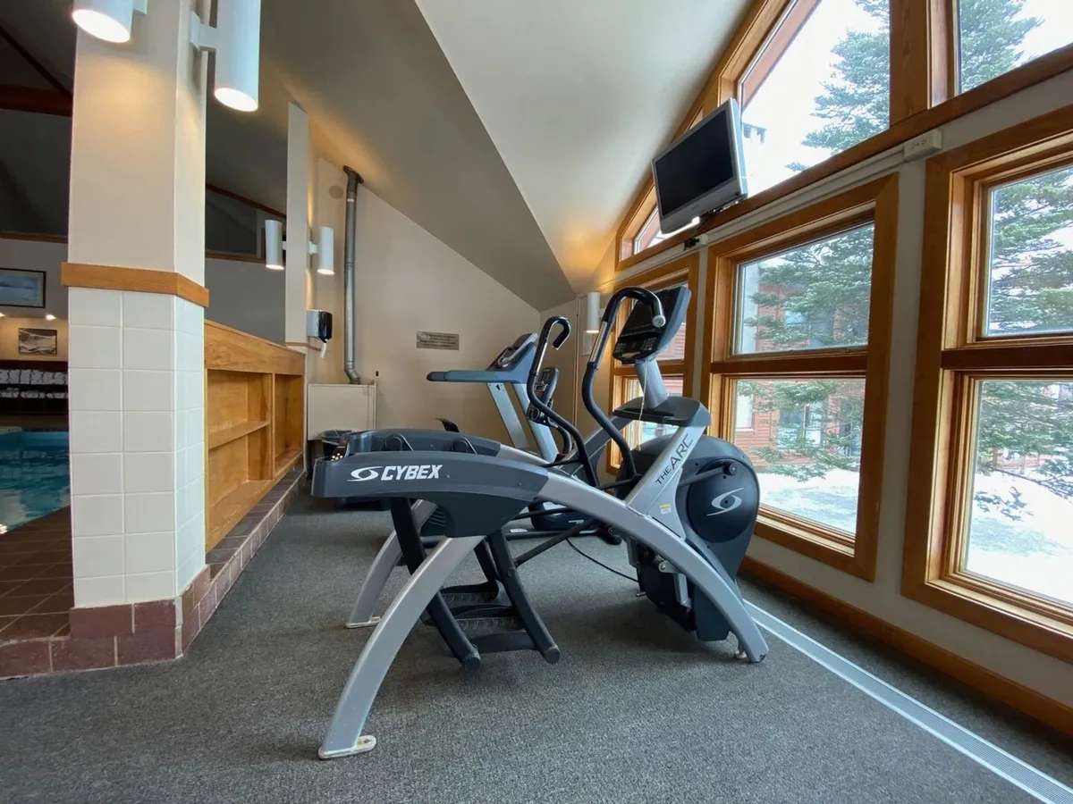 A modern gym space featuring elliptical machines positioned before large windows, accented by natural wooden details. The view outside, coupled with the well-equipped space, creates a motivational and refreshing backdrop for workouts.
