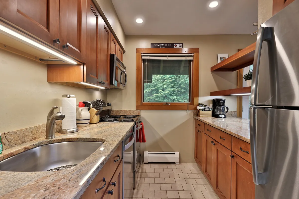 This cozy kitchen scene features a granite countertop, wood cabinetry, and a window framing a scenic mountain view. Kitchen appliances are visible and accessible, while hints of activity—such as baking cookies or preparing brunch—suggest the space's readiness for both casual and gourmet cooking. The layout is open, fostering entertaining and family gatherings.