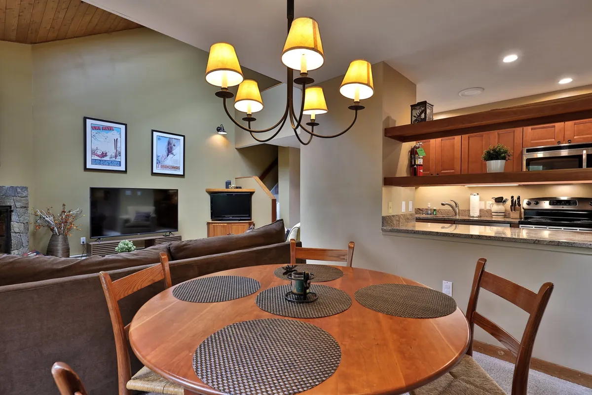 The image presents an integrated view of the dining table with a chandelier overhead, a comfortable sofa, kitchen area with appliances, and a television in the background. This open-plan interior layout allows easy movement and sightlines between the kitchen, dining, and living areas, making it perfect for communal dining and conversation.