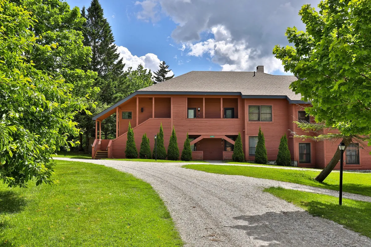 An inviting exterior view of the condo’s pink facade, set against a well-manicured lawn and bordered by mature trees. The gravel driveway leads guests to the entrance, hinting at serene surroundings and opportunities for outdoor enjoyment.