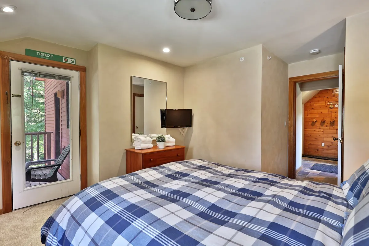 A cozy bedroom scene featuring a bed with plaid bedding, a wooden dresser, television, mirror, and a door leading to a balcony. The room is comfortable and homey, with a connection to the outdoors for added relaxation.