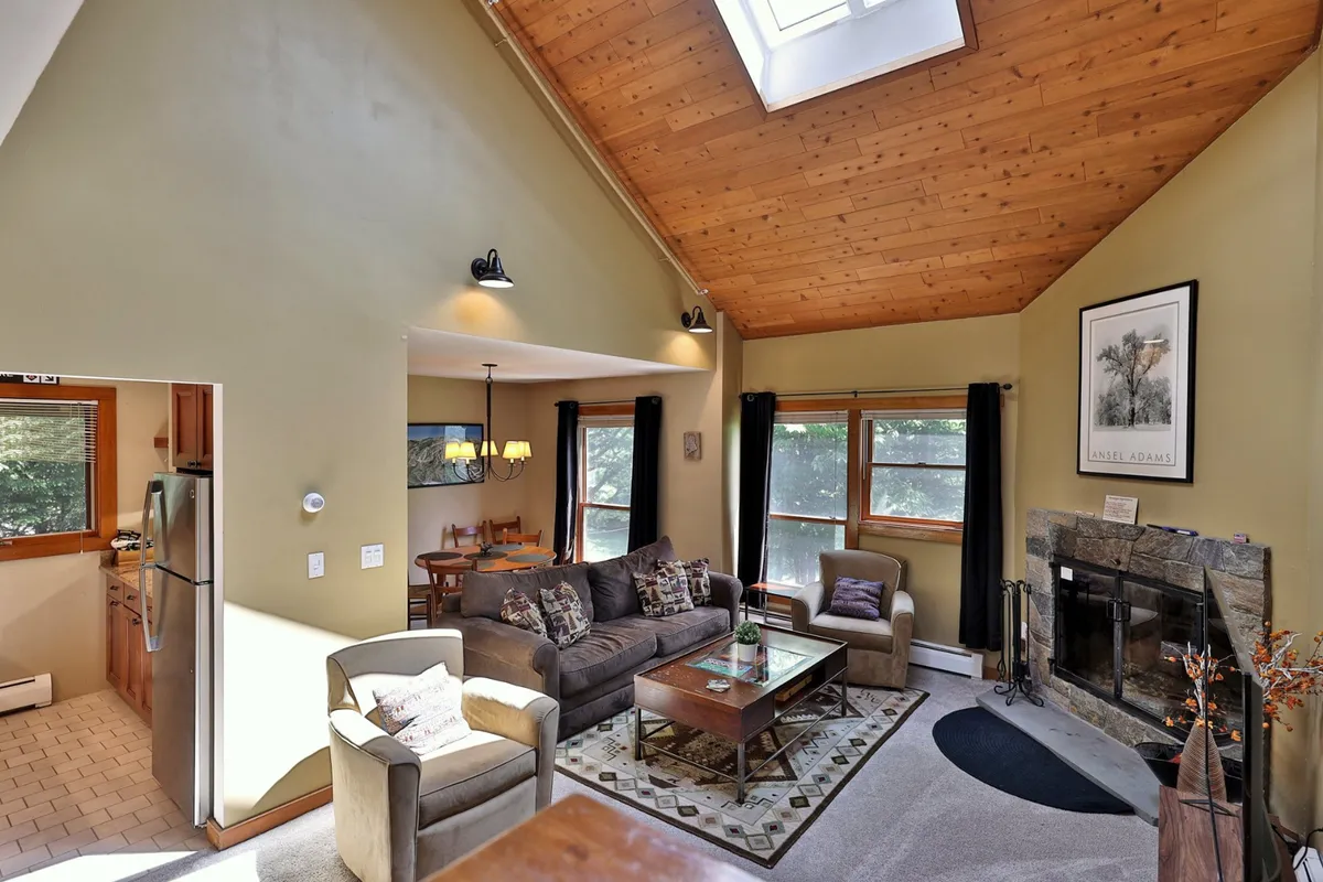 An inviting open-concept living and dining area, with a crackling fireplace, skylight overhead, comfortable seating, and a dining table for six. The space is bright and airy, perfect for family gatherings, game nights, or enjoying meals together in a serene mountain setting.