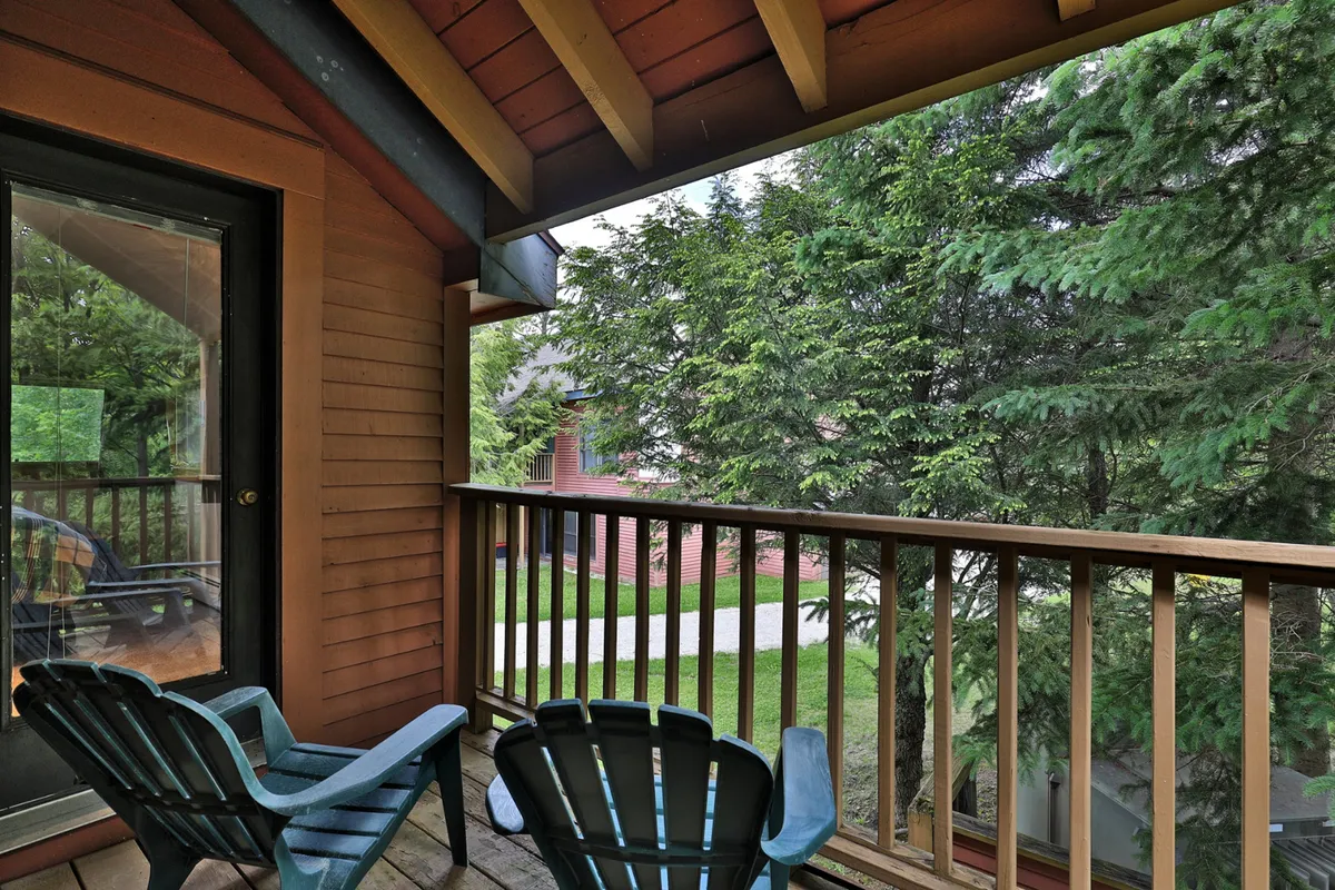 A peaceful exterior scene featuring a set of Adirondack chairs placed on a wooden deck or balcony, surrounded by lush natural greenery. The wooden railing hints at a private spot connected to the outdoors, perfect for watching wildlife, enjoying sunset views, or basking in the fresh Vermont air.