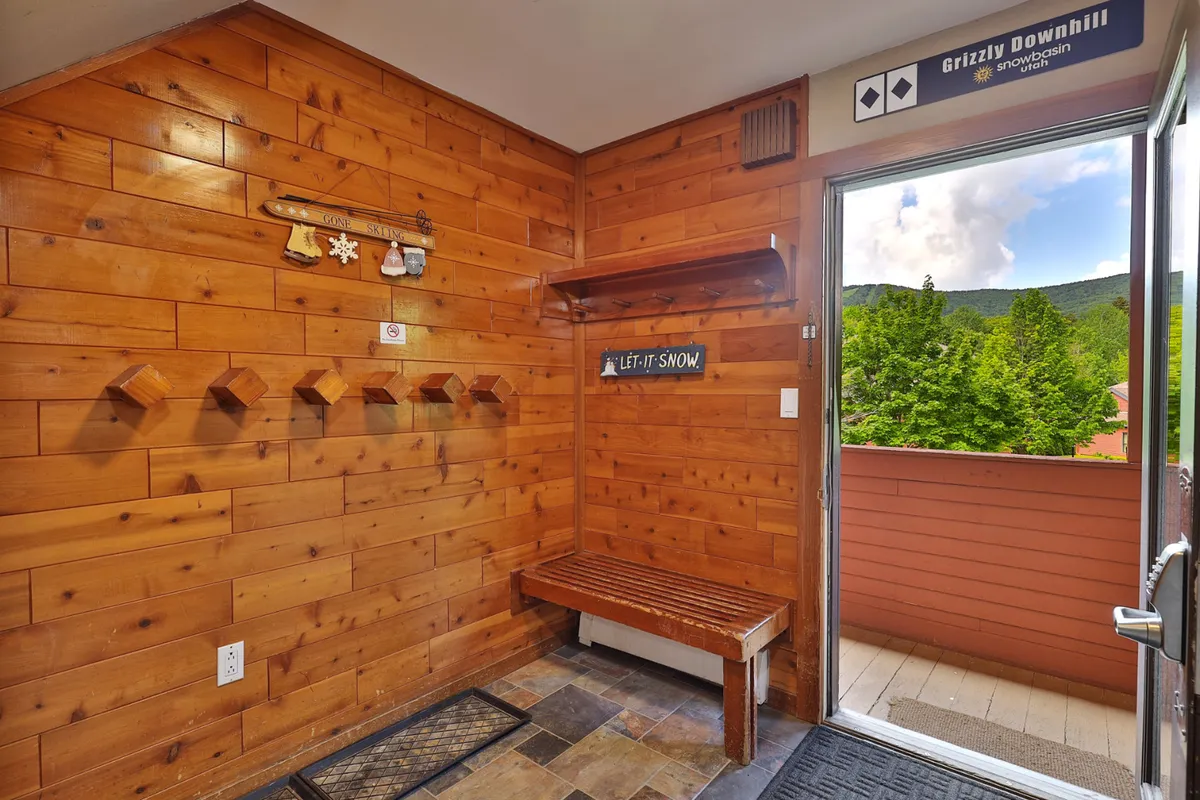 A cozy mudroom featuring warm wooden walls, a built-in bench, shelves with hooks for coats and gear, and welcoming signs. The space is clearly designed for guests to prepare for outdoor adventures or return from them, serving as a transition area between inside comfort and mountain activities.