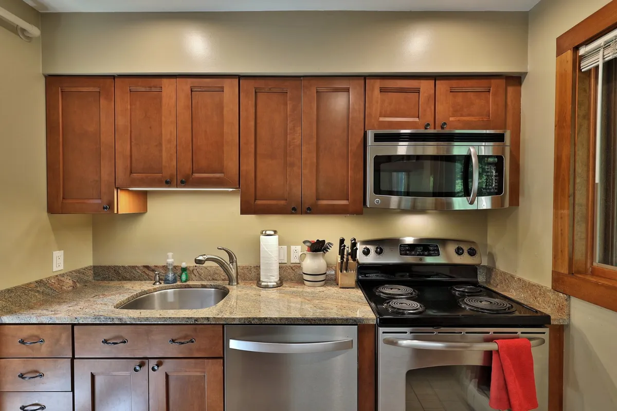 The image displays a spacious kitchen with elegant kitchen cabinetry, gleaming stainless steel appliances, and a polished granite countertop. Sunlight streams through the window, giving the space a warm, inviting atmosphere. The setup clearly emphasizes functionality as well as aesthetic appeal, with ample surfaces for meal prep and an open design ideal for group cooking or casual gatherings.