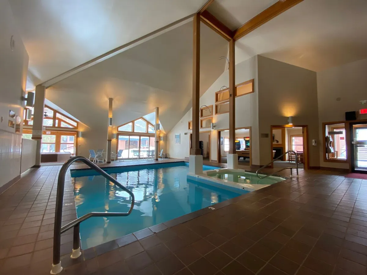 A spacious indoor swimming pool area featuring both a large pool and an adjacent jacuzzi. Sunlight pours in through oversized windows, illuminating wooden beams overhead and creating a warm, inviting ambiance. The design blends relaxation and social spaces, perfect for both energetic swims and tranquil lounging.