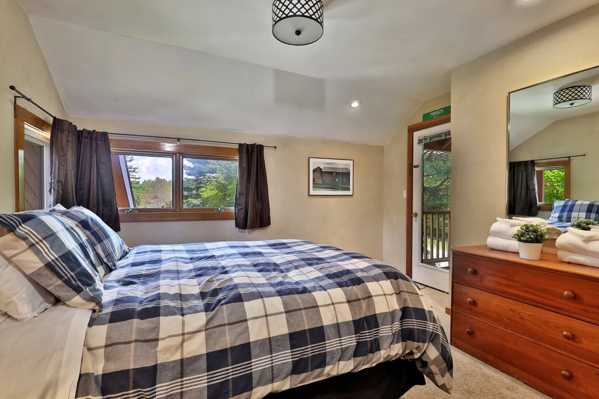 This image captures a serene bedroom with plaid bedding, a window offering views of nature, and a dressing table with mirror. The inviting atmosphere emphasizes comfort and relaxation, aligning well with the primary bedroom's description.